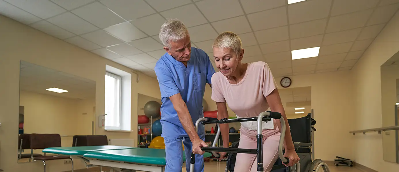 Frau übt mit Pfleger das Gehen an Gehhilfe.
