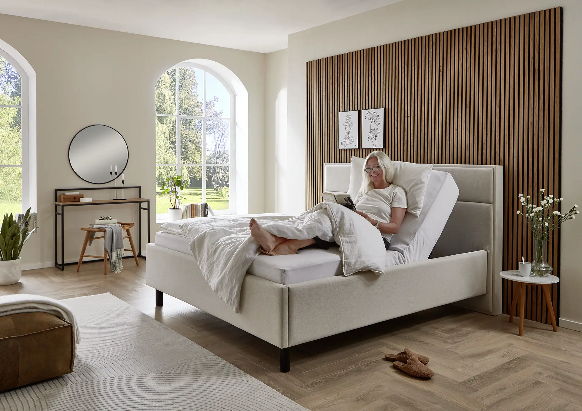 Beiges Lift Up Polsterbett in einem hellen, modern eingerichteten Schlafzimmer; eine Frau sitzt bequem in der hochgefahrenen Rückenlehne – höhenverstellbares Komfort- und Pflegebett.